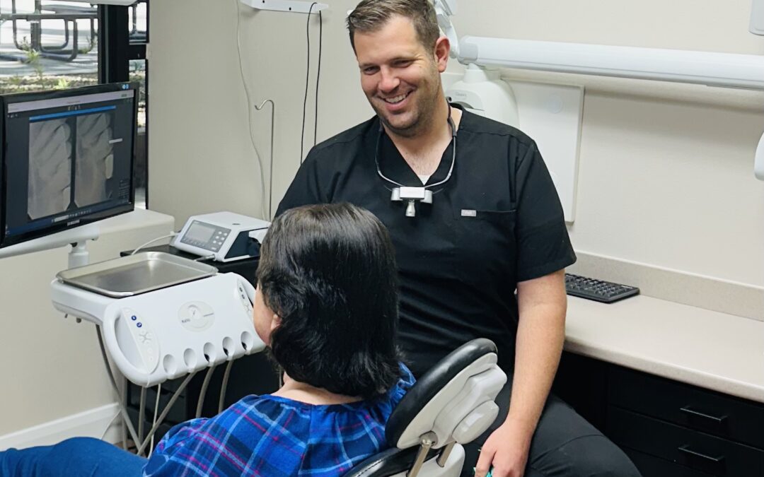 A dentist in a black uniform is smiling and conversing with a patient seated in a dental chair, who is turned towards him, listening attentively. The room is equipped with modern dental technology, including a monitor displaying x-rays to the left. The atmosphere is friendly and professional, highlighting a positive patient-dentist interaction